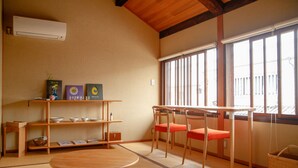 A traditional Japanese room with tatami flooring, a wooden table, two chairs, a shelf with bowls, and a window with vertical blinds. - Temari Hotel Yukikai (Kurashiki)