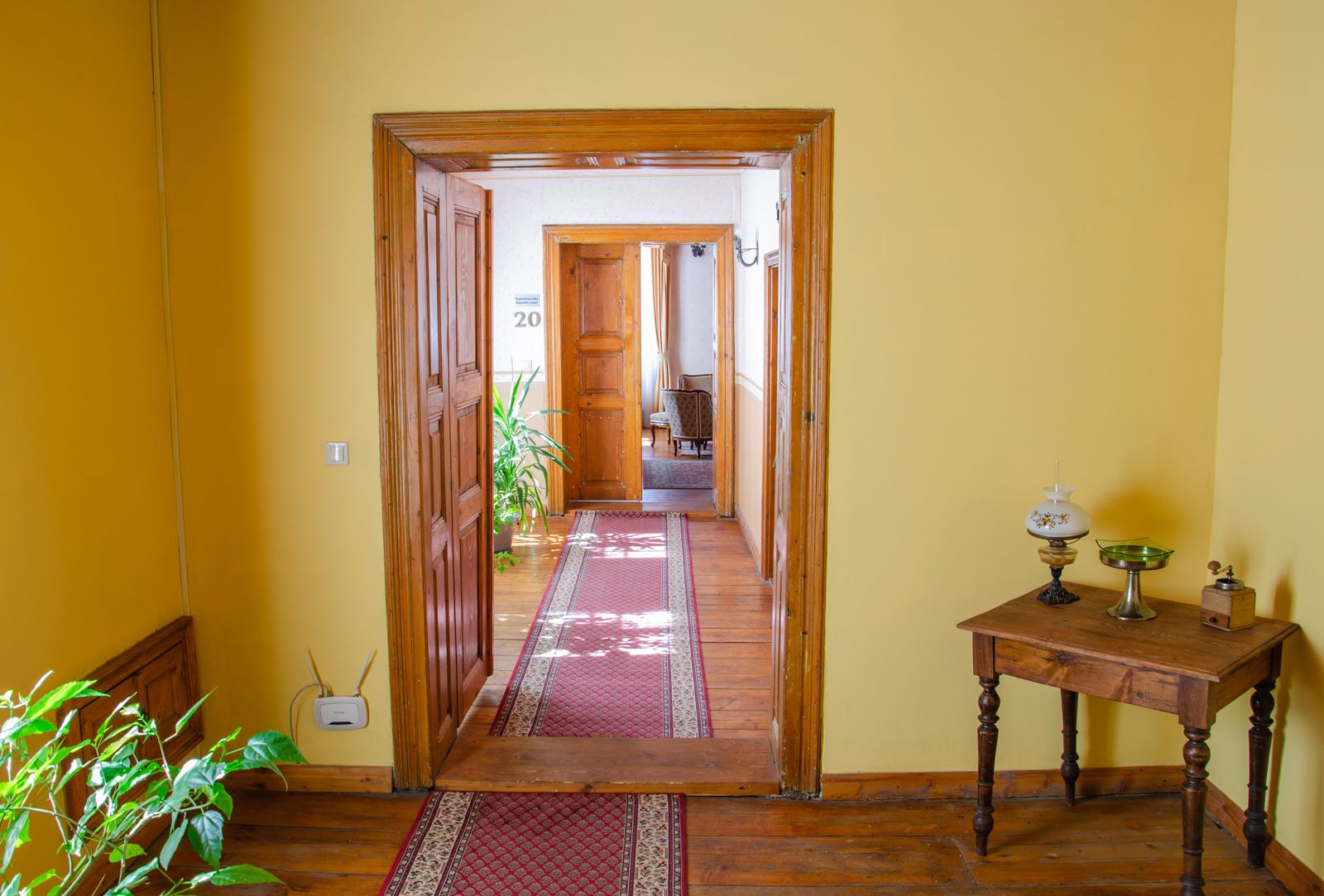 Un couloir avec des portes en bois, un tapis rouge à motifs et une petite table ornée d’objets décoratifs.