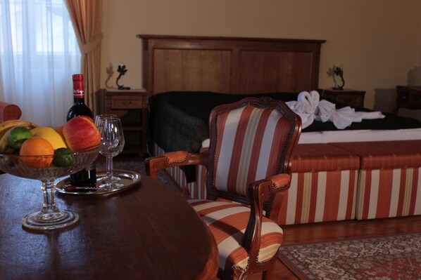 A hotel room with a wooden bed, a striped armchair, a dining table with fruit and wine glasses, and a nightstand with a bottle.