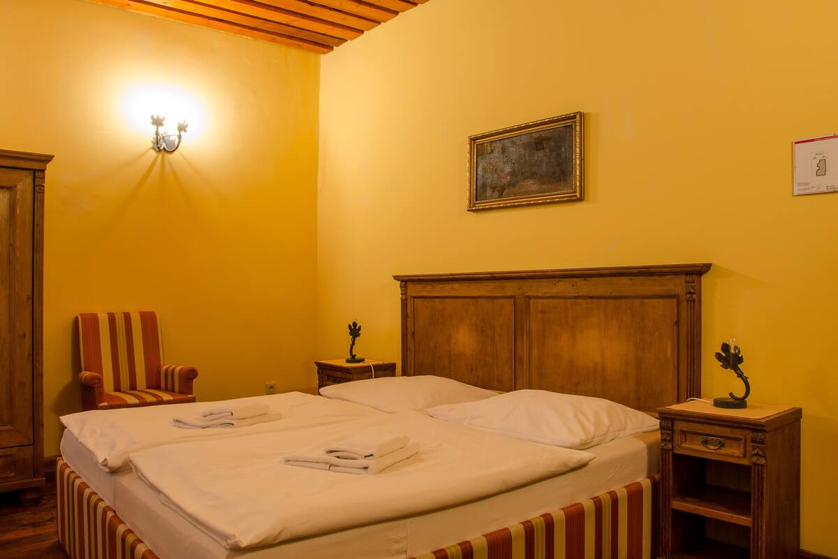 A hotel room with a double bed, a wooden headboard, a striped armchair, a nightstand with a lamp, and a painting on the wall.