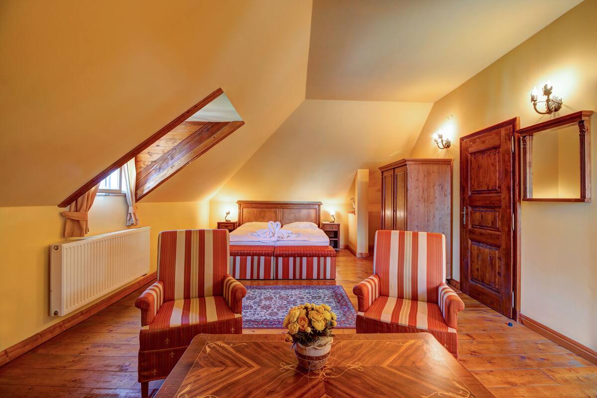 A bedroom with a sloped ceiling, a bed with a striped bedspread, two striped armchairs, a wooden table with a vase of flowers, a radiator, and a wooden wardrobe.