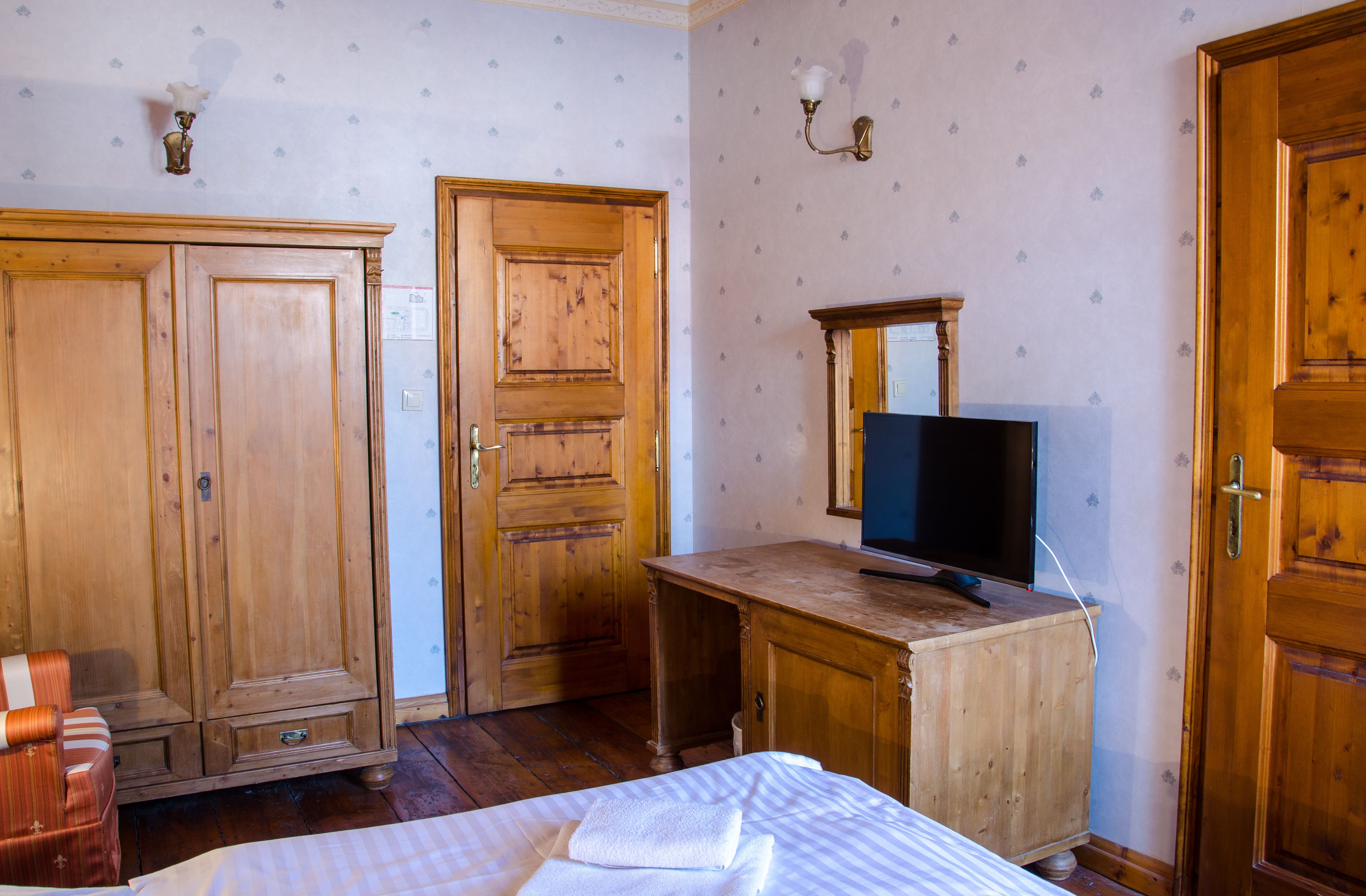 A room with wooden wardrobes, a bed with a white and blue striped bedspread, a wooden desk with a television, and a wall-mounted light fixture.