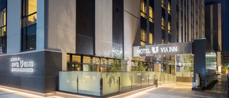 A modern hotel exterior with illuminated signage, a glass entrance, and a tiled pavement.