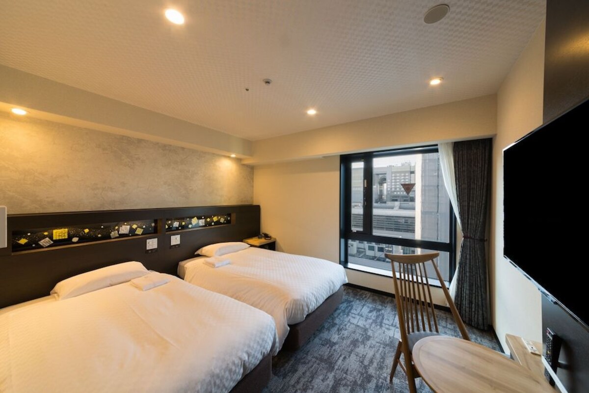 A hotel room with two beds, a desk, a chair, a TV, and a large window.