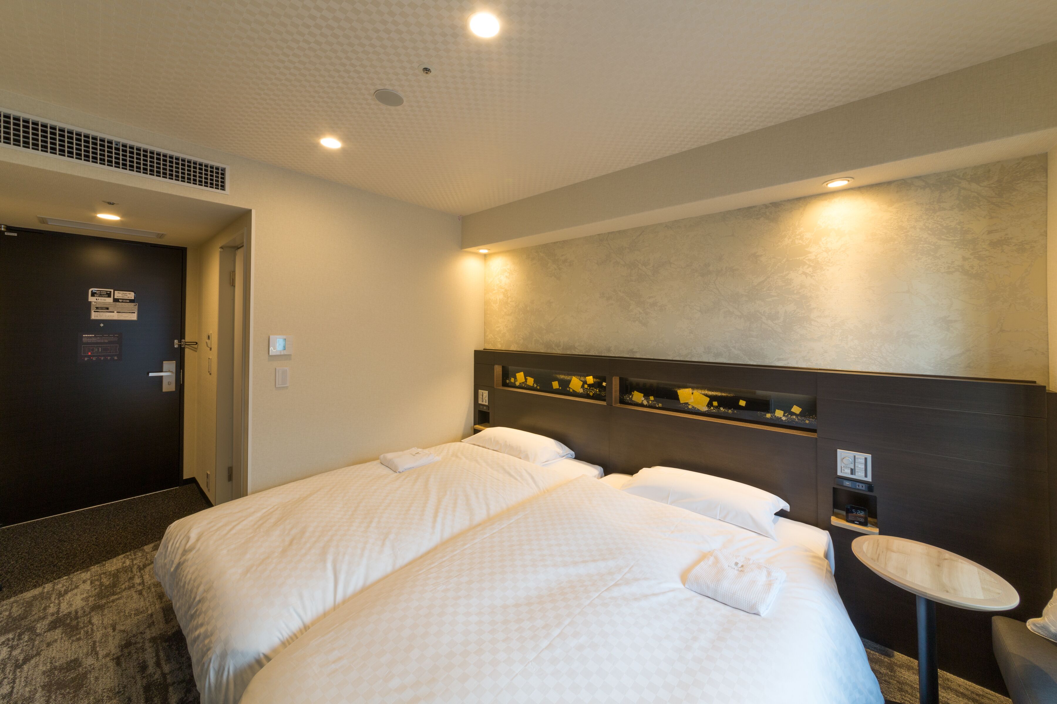 Ett hotellrum med två sängar, ett litet runt bord och en vägghylla med dekorativa föremål.