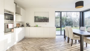 A modern kitchen with white cabinetry, a herringbone wood floor, a dining area with a dark table and chairs, and a large window with a view of trees. - Killester Self Catering Cottage (Dublin)