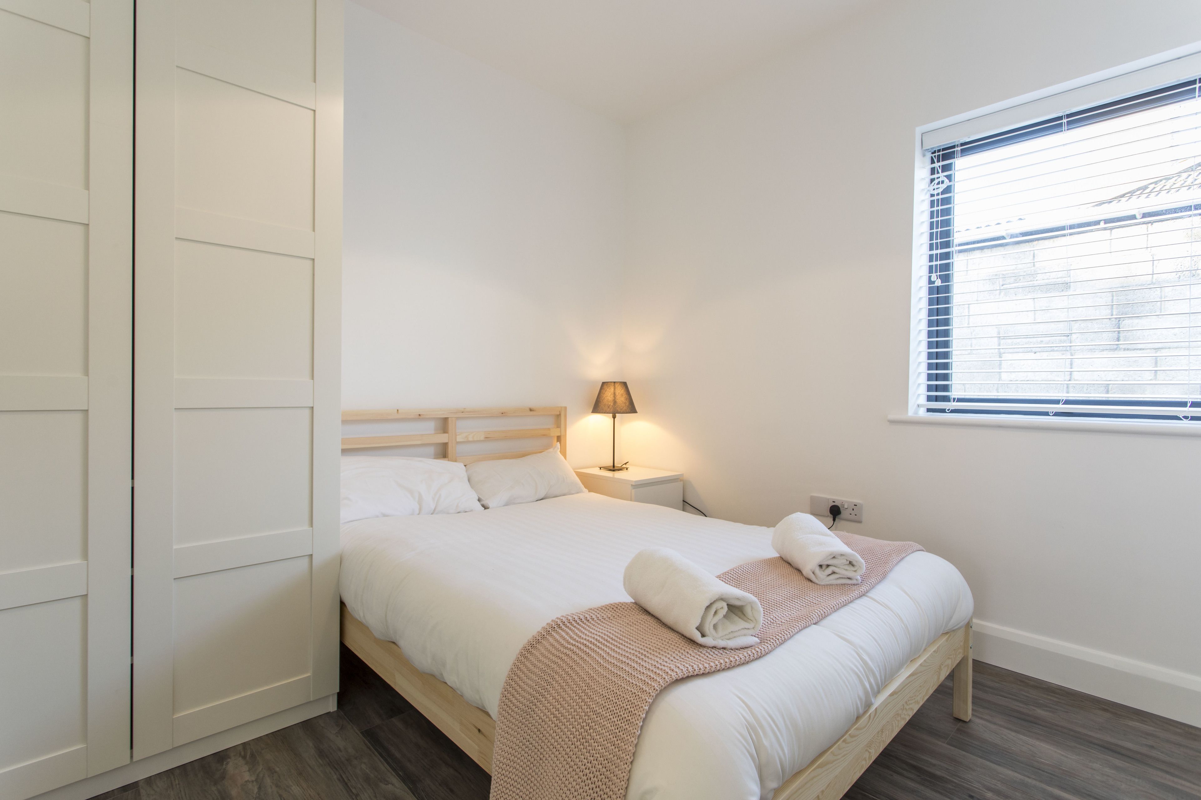 Una cama bien hecha con cabecera de madera, una lámpara de noche y una ventana con persianas.