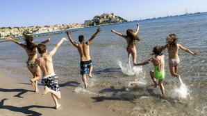 A beach scene with people jumping into the water, a clear sky, and a distant view of a fortified building. - Village Club Miléade Calvi (Calvi)