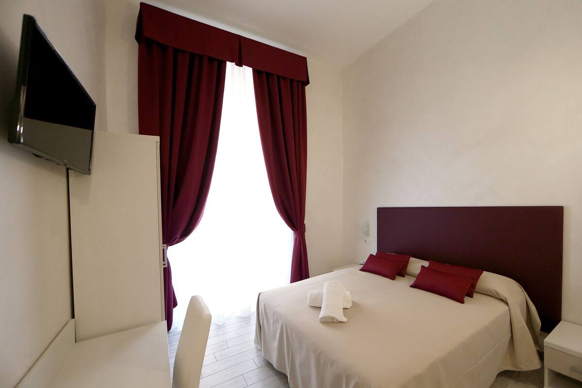 A hotel room with a bed, a TV mounted on the wall, and a window with red curtains.
