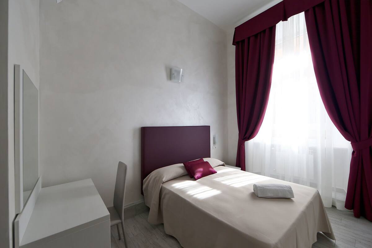 A modern hotel room with a bed, a desk, and a large window with red curtains.