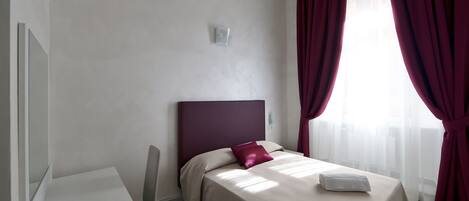 Moderni hotellihuone, jossa on sänky, työpöytä ja suuri ikkuna, jossa on punaiset verhot.