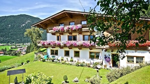 Una cabaña de dos pisos con balcones decorados con flores, rodeada de exuberante vegetación y montañas al fondo.