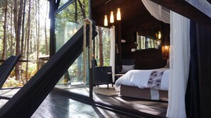 Une chambre dotée d’un grand lit, d’un coin salon avec des fauteuils et offrant une vue sur la forêt grâce à de grandes fenêtres.