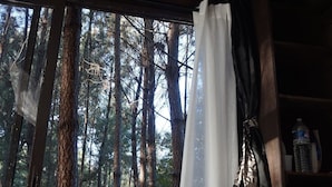 Una habitación acogedora con techo de madera, una silla de mimbre, una mesita y vistas a un bosque a través de una ventana con cortinas blancas.