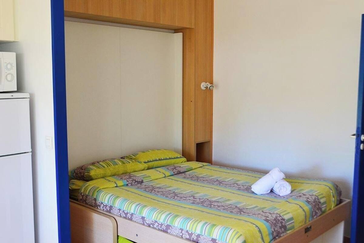 Ein kleines Zimmer mit einem Einzelbett, einem Holz-Kopfteil, einer weißen Wand und einem kleinen, weißen Gerät links.