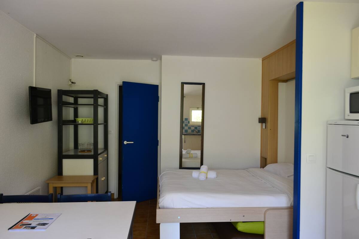 Ein kleines, sauberes Zimmer mit einem Bett, einem Schreibtisch, einem Stuhl, einem Spiegel und einem Fernseher.