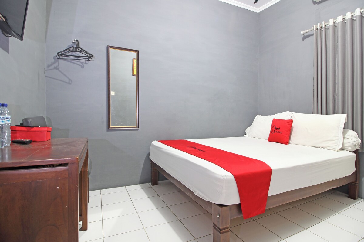 A hotel room with a bed, a wooden desk, a mirror, and a red and white bedspread.