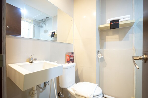A bathroom with a white sink, toilet, and shower. There is a mirror above the sink and a shelf with towels above the toilet. - RedDoorz Apartment @ Bogor Valley (Bogor)
