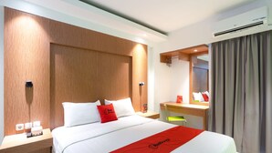 A hotel room with a bed, a desk, a chair, a mirror, and an air conditioning unit. - RedDoorz Apartment @ Bogor Valley (Bogor)