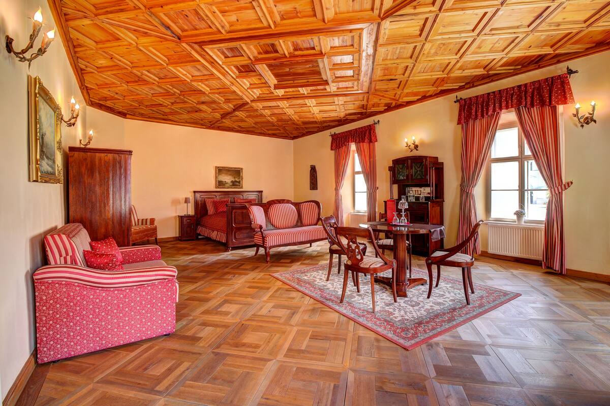 A cozy living room with a wooden ceiling, a sofa, chairs, a dining table, and a patterned rug.