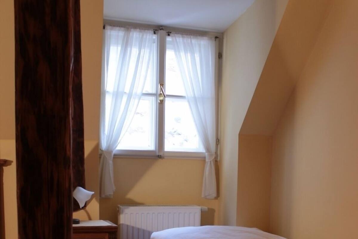 A small, cozy bedroom with a single bed, a wooden nightstand, a window with sheer curtains, and a radiator.