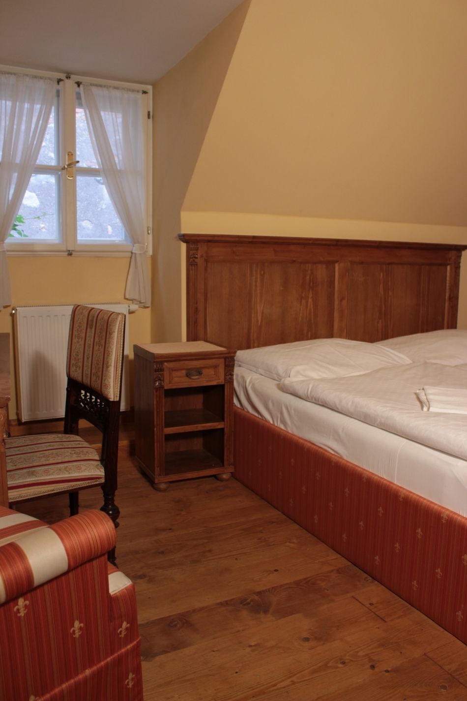 Un dormitorio con cabecera de madera, una cama individual, una mesita de noche, una silla y una ventana con cortinas.