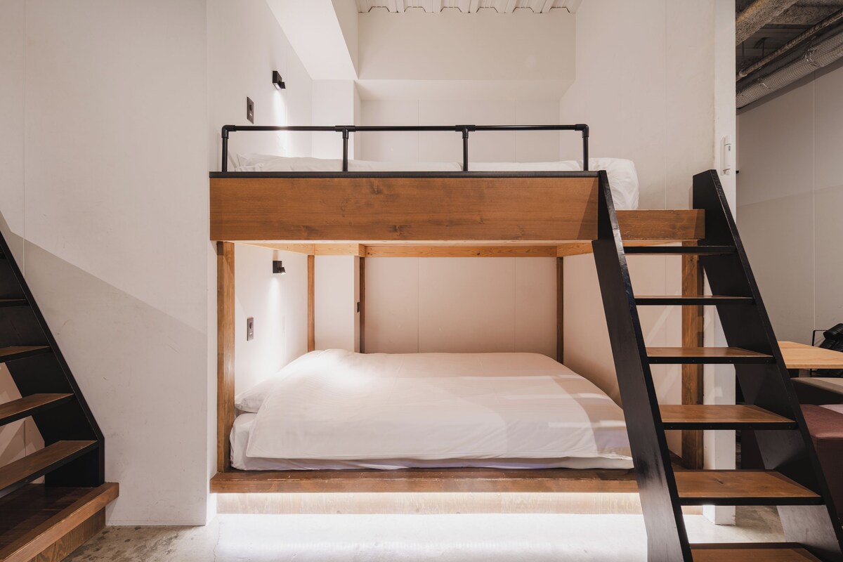 Un lit superposé avec une structure en bois et une literie blanche, un escalier sur le côté gauche et un mur blanc.