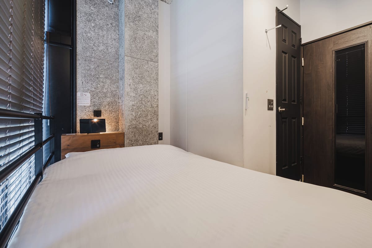Une chambre à coucher avec un lit, une table de chevet en bois, un mur d’accent en pierre, une porte sombre et une fenêtre avec des stores.