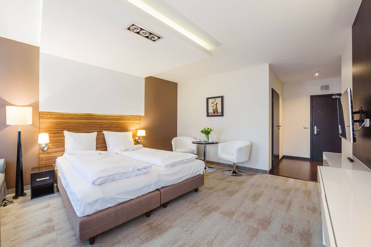 Habitación de hotel con una cama grande, lámparas de noche, una mesita con un florero, una silla y un televisor.