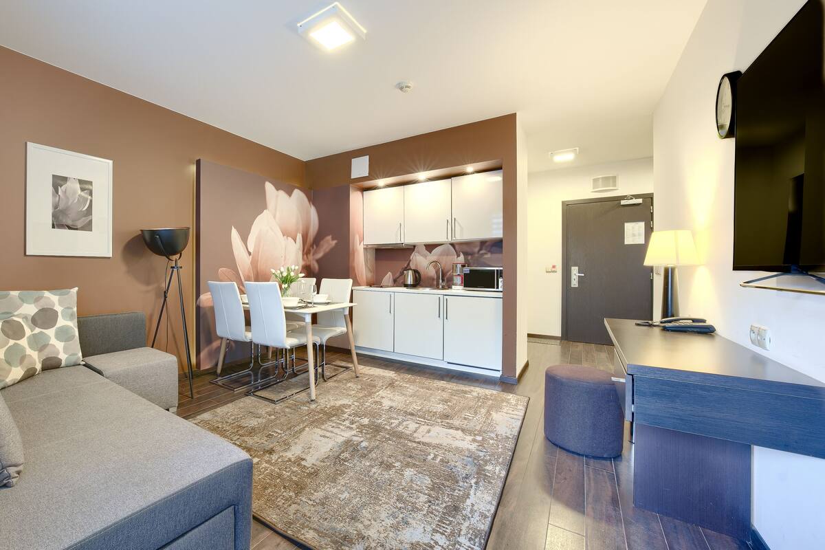 Una sala de estar moderna con un sofá, un comedor con mesa y sillas, una cocina con armarios blancos y un televisor montado en la pared.