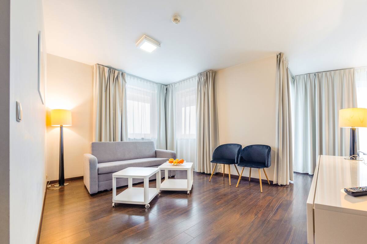 Una habitación de hotel moderna con un sofá gris, dos sillones, una mesita blanca con fruta, un televisor y un control remoto.