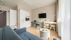 A modern living room with a sofa, a dining area with chairs, a kitchen area with cabinets, a flat-screen TV mounted on the wall, and a small round table with a cup and a book.