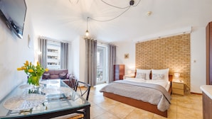 Une chambre d’hôtel moderne avec une table à manger en verre, un canapé, un lit avec un mur décoratif en briques et une vue sur la ville.