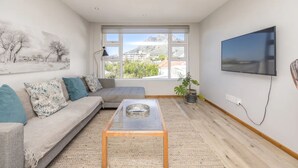 Ein modernes Wohnzimmer mit einer Couch, einem Glastisch, einem an der Wand montierten Fernseher und einem großen Fenster mit Blick auf Gebäude und Berge.