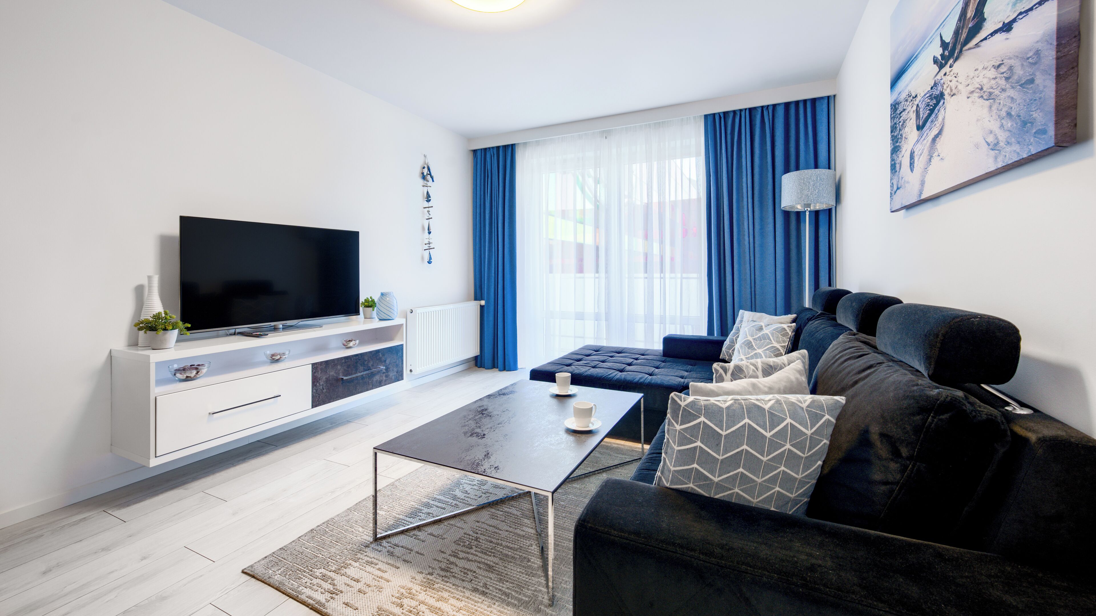 A modern living room with a flat-screen TV, a sofa, a coffee table, and a large window with curtains.