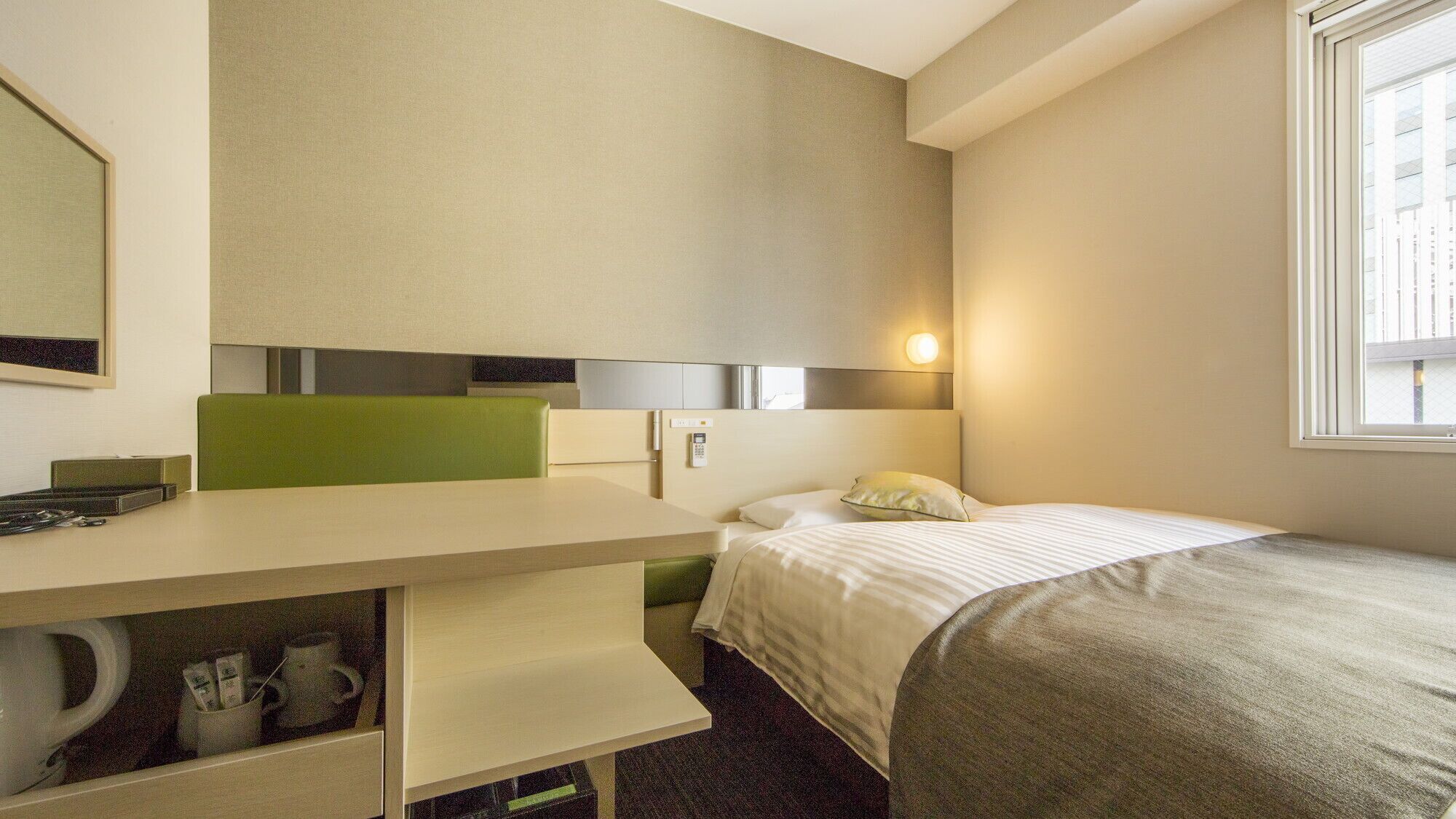 Ett hotellrum med en säng, ett skrivbord med en telefon, en spegel och ett fönster.