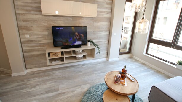 A modern living room with a flat-screen TV, wooden flooring, a round coffee table, and a large window with natural light. - Apartments Angelov (Varna)