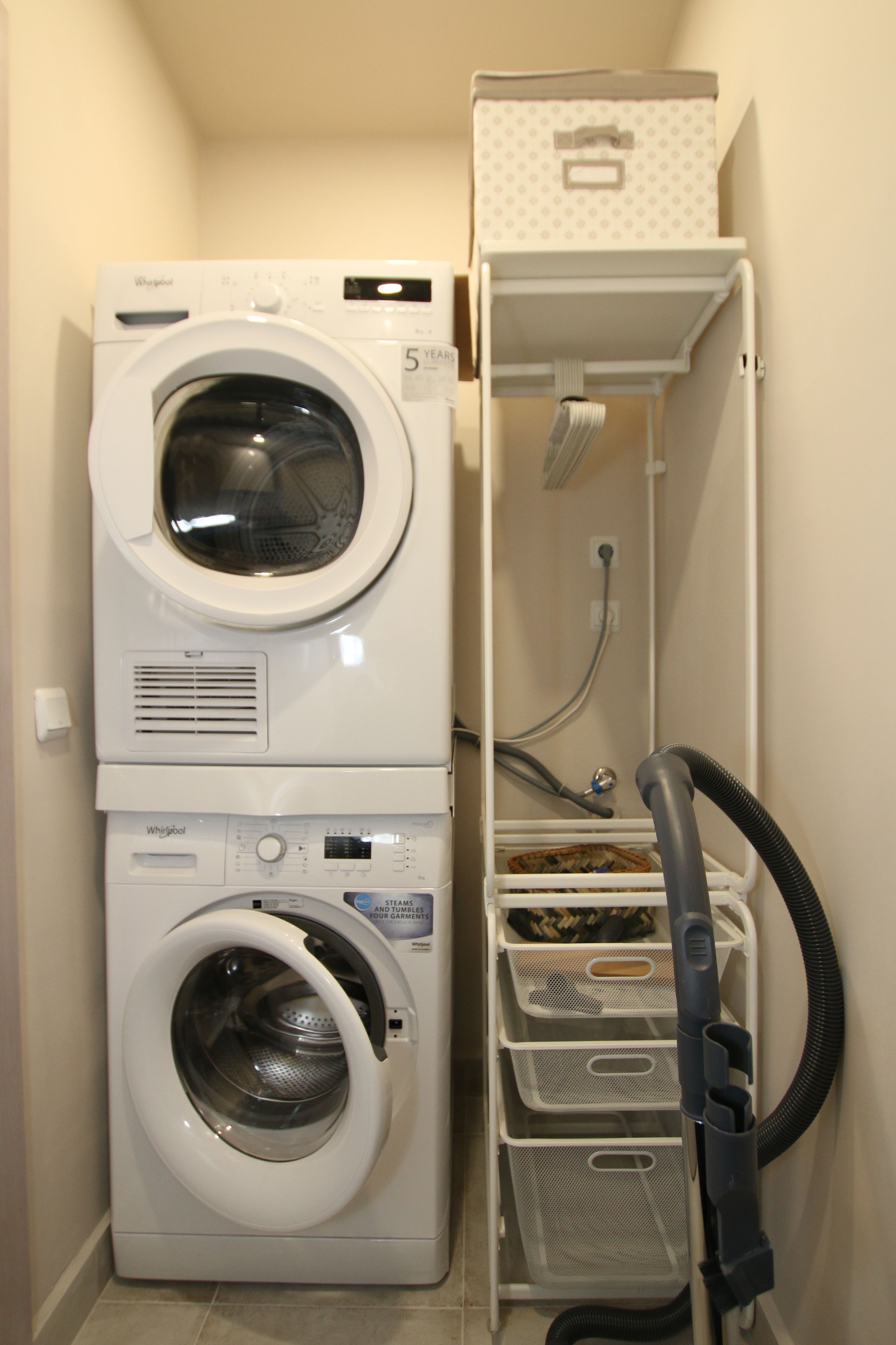 A compact laundry area with a front-loading washer and dryer, a drying rack, and storage bins.