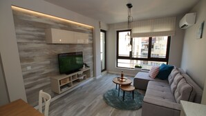 A modern living room with a flat-screen TV, a sofa, a small table, and a window with a view of buildings. - Apartments Angelov (Varna)