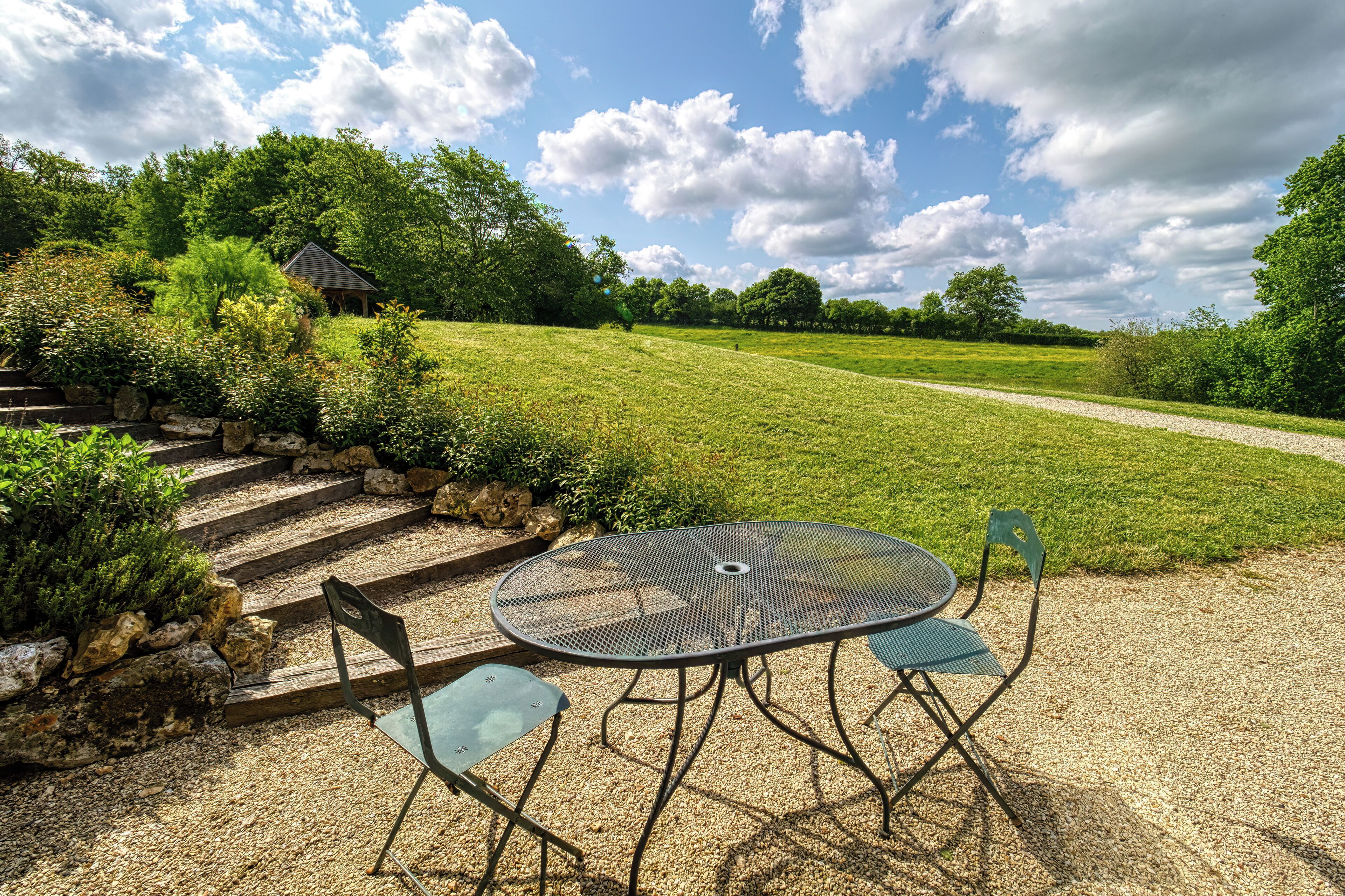 A patio with a round metal table and two chairs, surrounded by a gravel area and a grassy field with a stone staircase leading up.