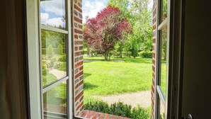 A view through a window with a brick exterior, revealing a grassy yard with trees and a clear sky. - Chambres d'hôtes - La Verrerie d' Ivoy (Ivoy le Pre)