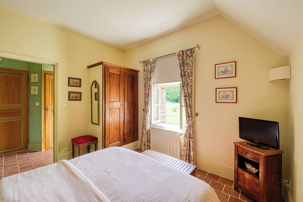 Une chambre à coucher avec un lit, une armoire en bois, une petite table avec un téléviseur et une fenêtre avec des rideaux.