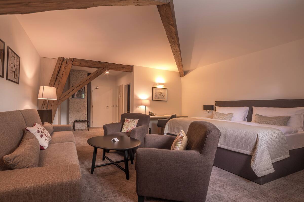 A hotel room with a bed, two sofas, a small table, and a desk. There is a staircase in the background.