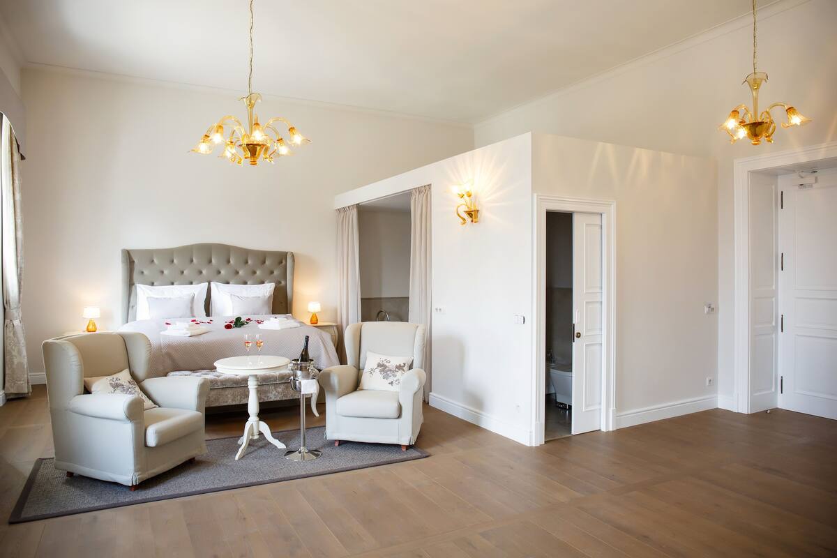 A bedroom with a large bed, two armchairs, a small table, and a chandelier.