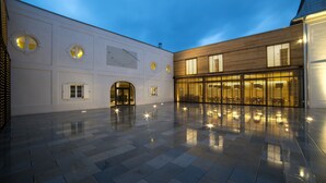 Un patio espacioso con suelo de baldosas reflectantes, un edificio de madera con ventanas amplias y un edificio blanco con ventanas circulares y una puerta.