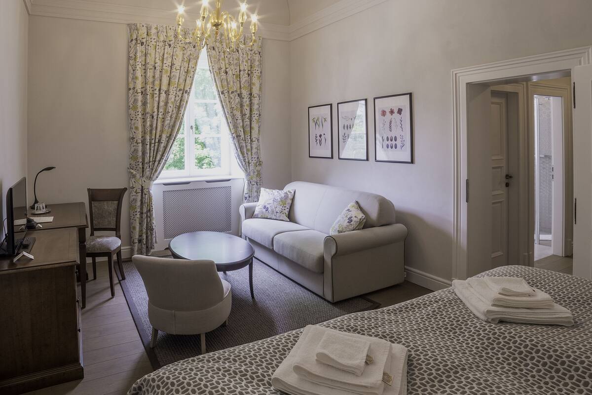 A bedroom with a bed, a sofa, a desk, and a chair. There is a window with curtains, a chandelier, and framed pictures on the wall.