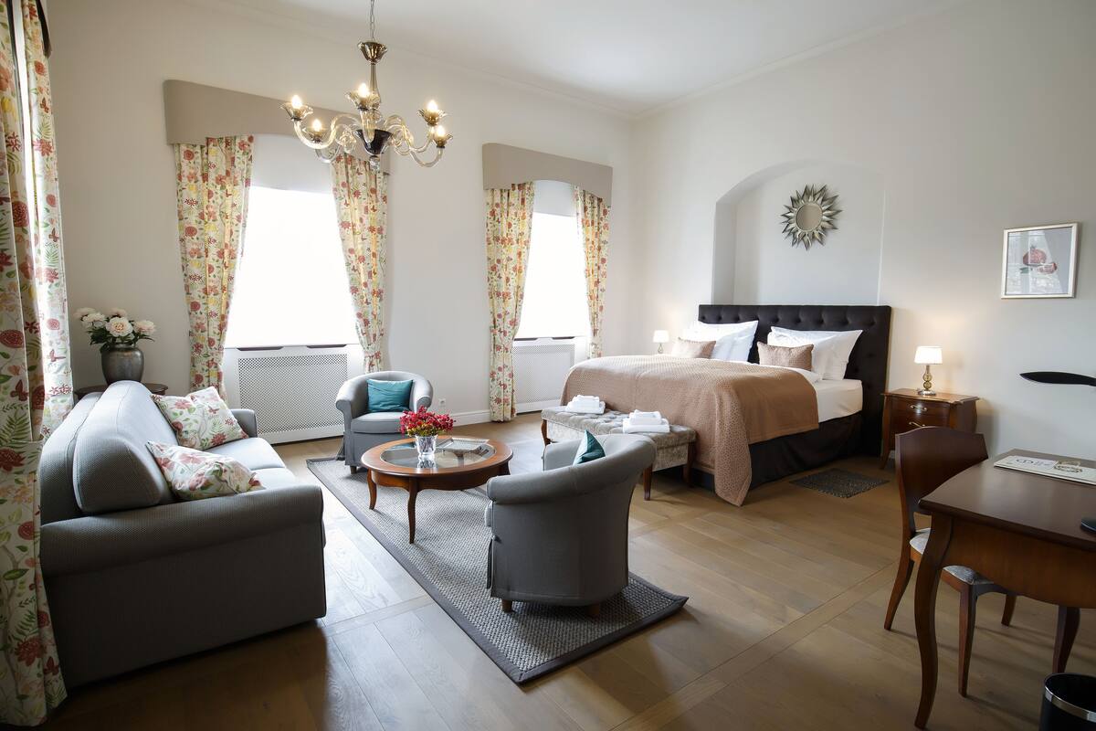 A modern hotel room with a bed, sofa, armchair, dining table, and a chandelier.