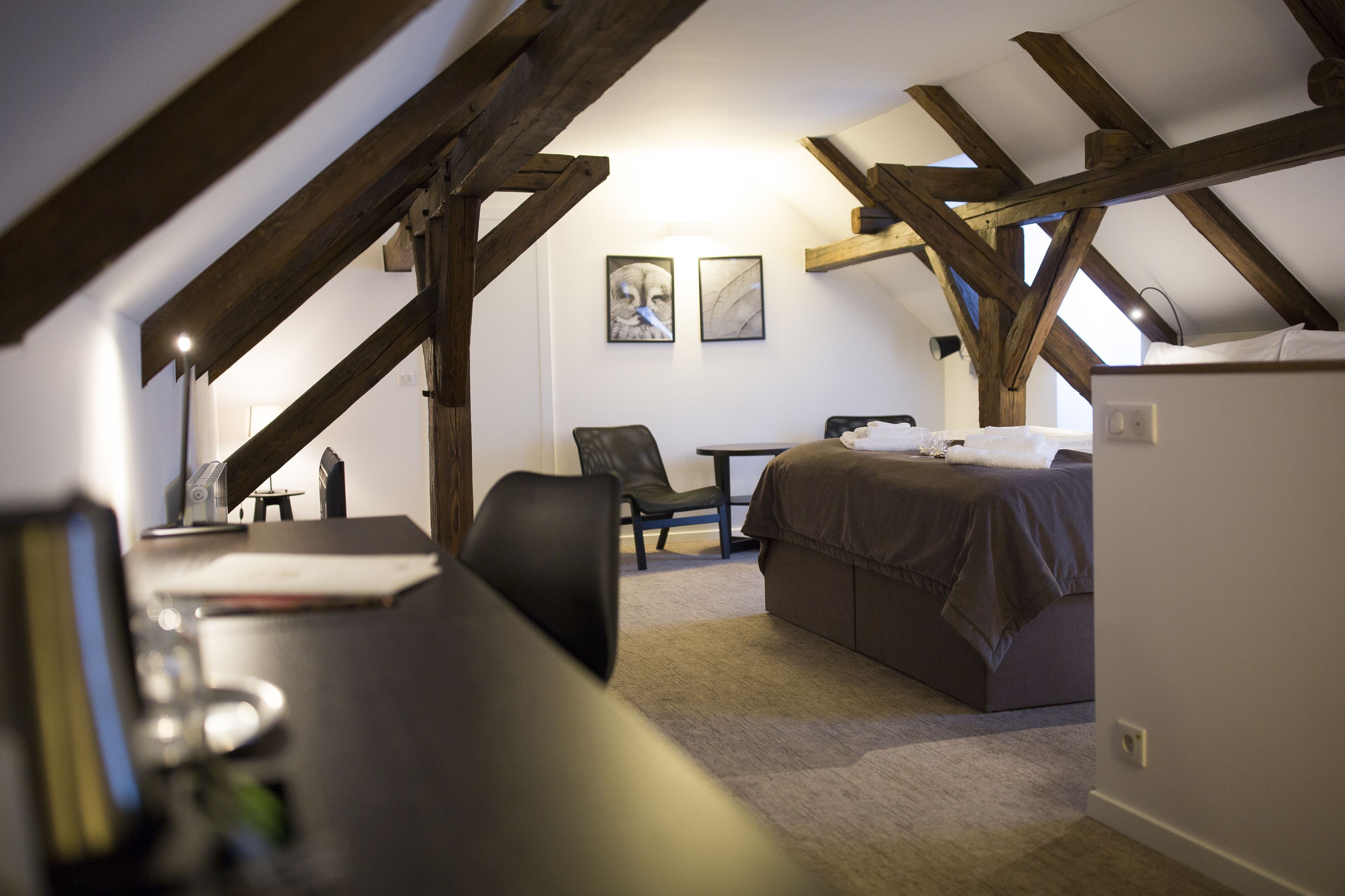 A bedroom with a bed, a chair, a desk, and a table. There are wooden beams on the ceiling and a picture on the wall.