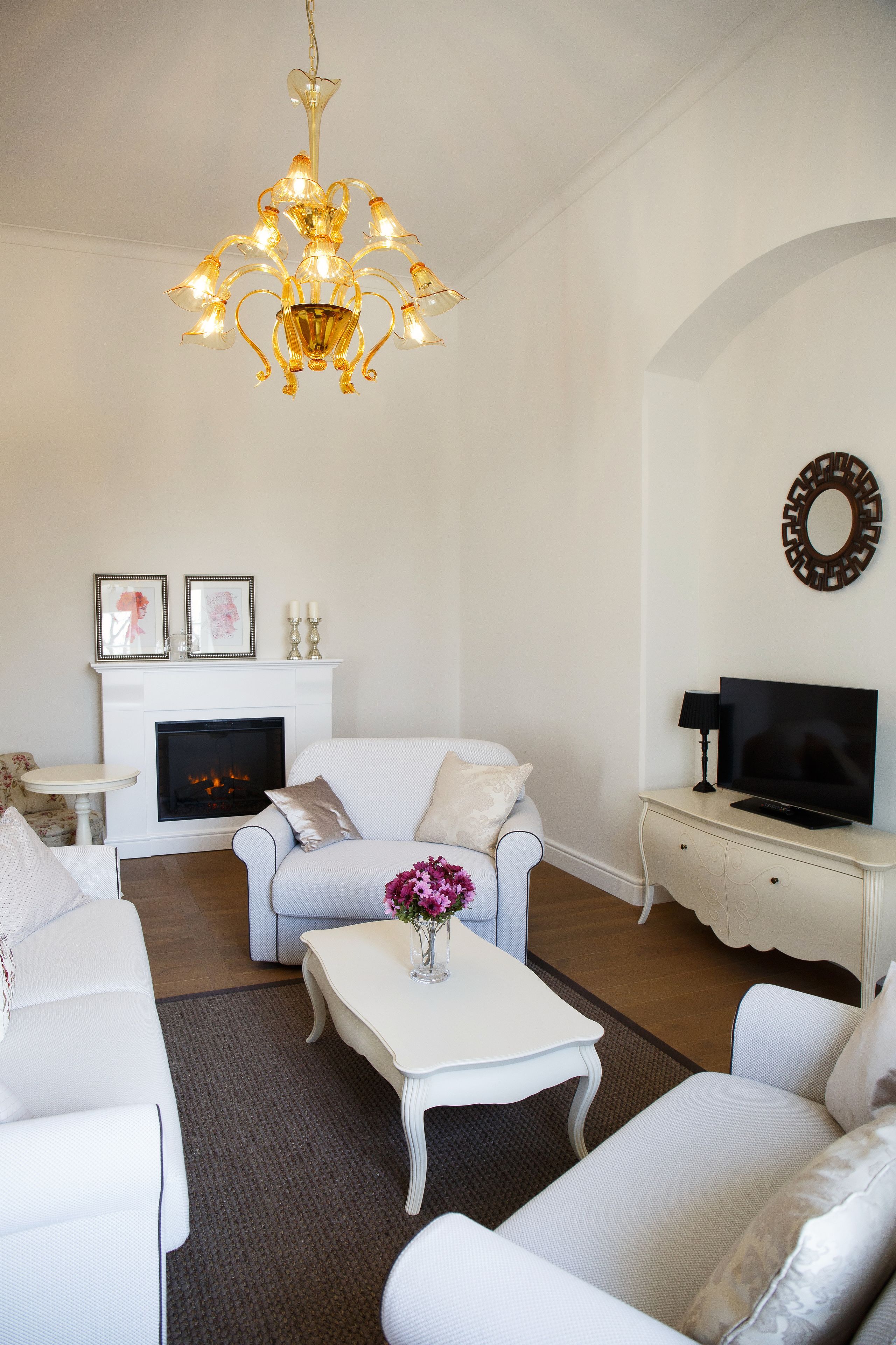 Una sala de estar con chimenea, sofás blancos, una mesa de centro con flores, un televisor sobre una mesita auxiliar y una araña.