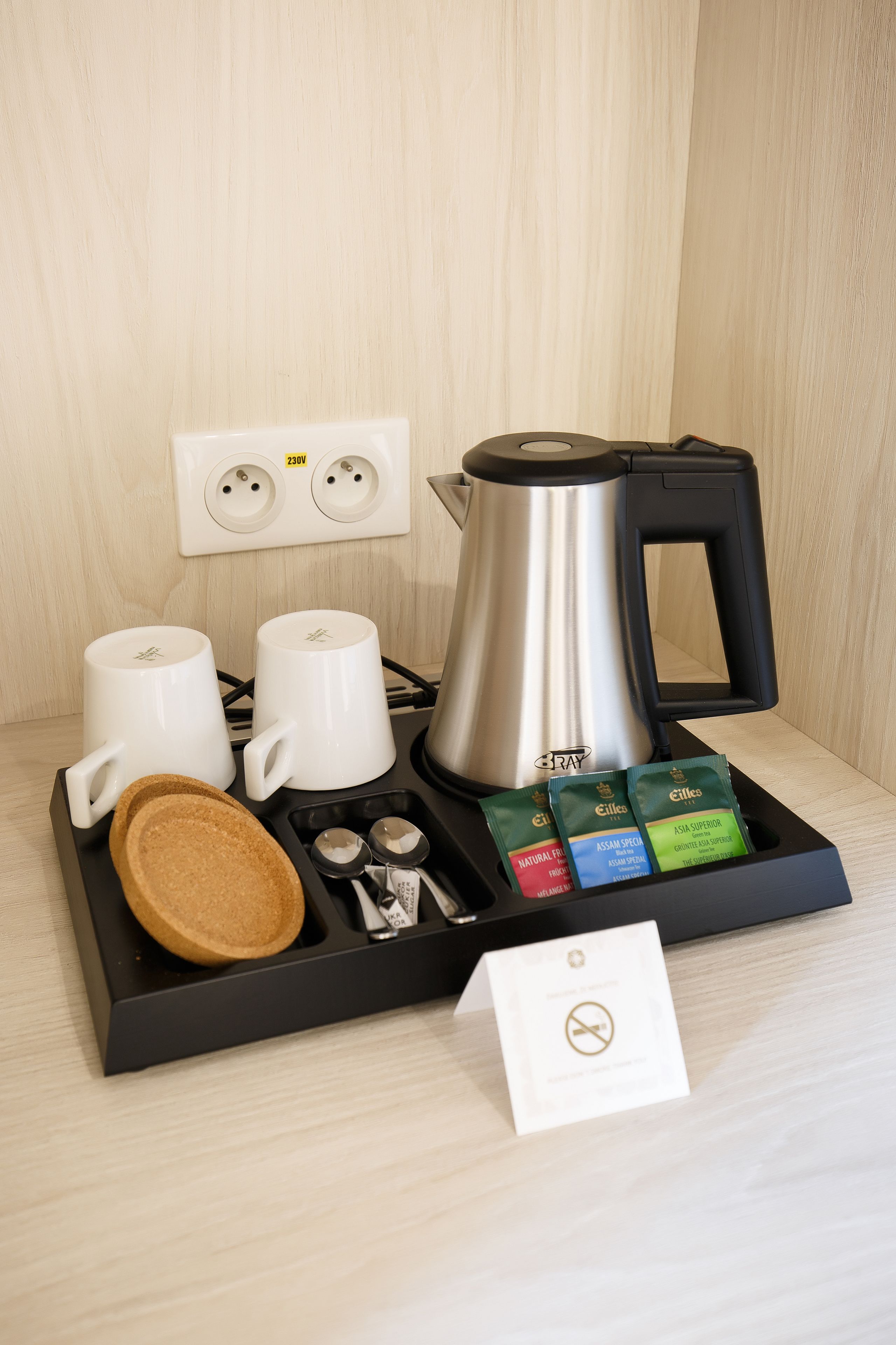 Un coin café comprenant une bouilloire, deux tasses blanches, un plateau avec des dosettes de café et un sachet de sucre.